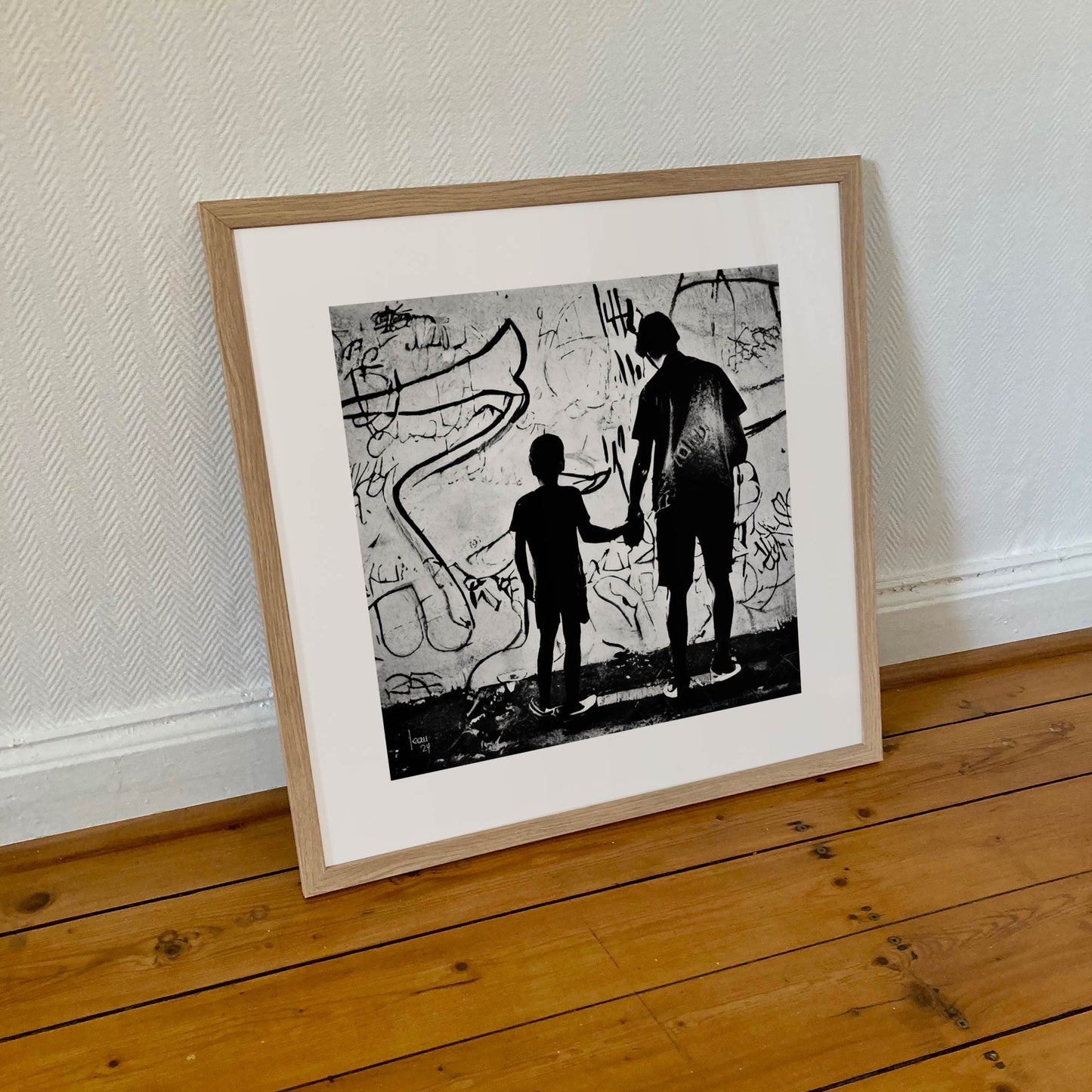 Œuvre "Paternité" encadrée au format 50×50 cm, silhouettes d’un père et de son enfant devant un mur tagué, encadrement minimaliste en bois clair.