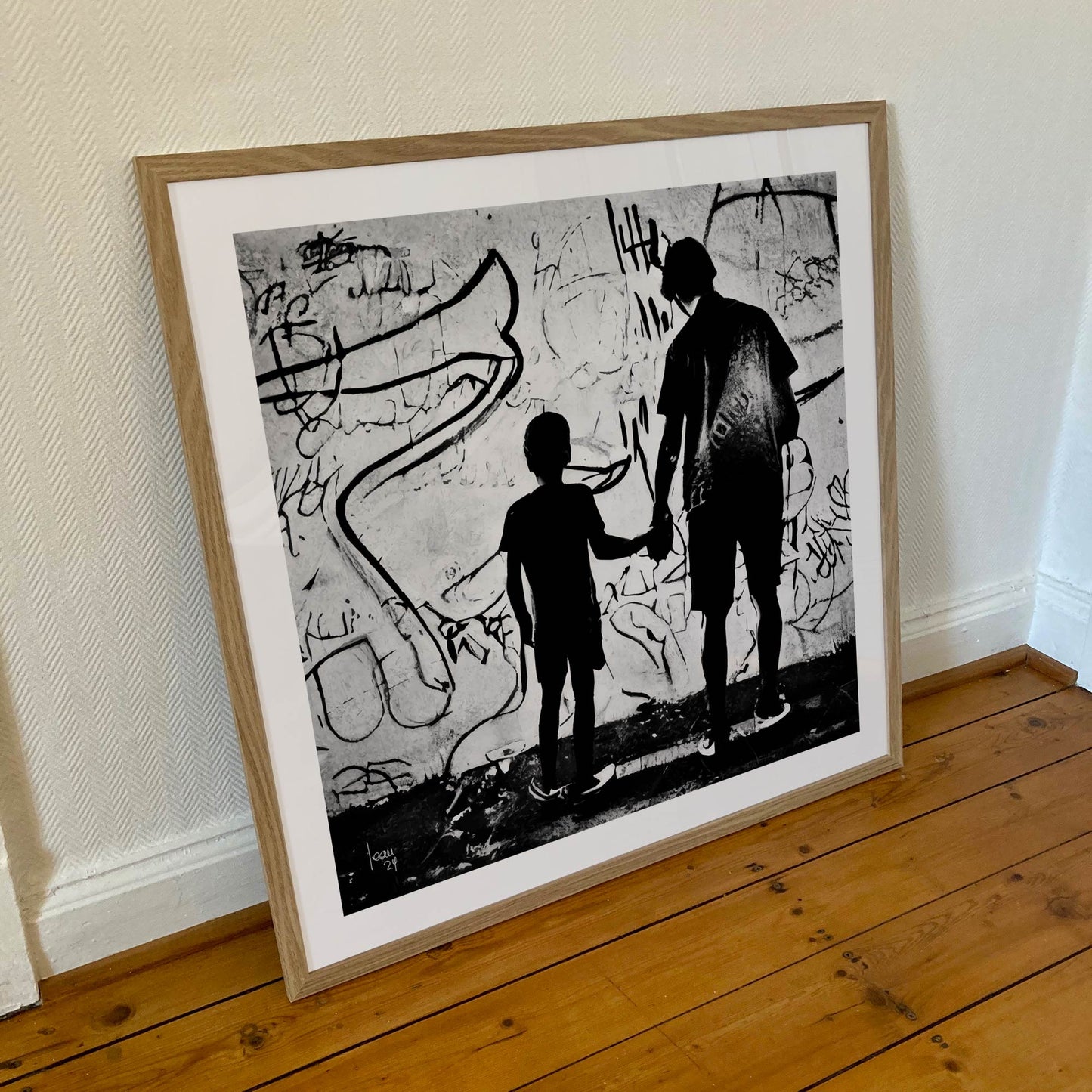 Œuvre "Paternité" encadrée au format 70×70 cm, duo de silhouettes en noir et blanc face à un mur artistique, encadrement sobre et élégant.