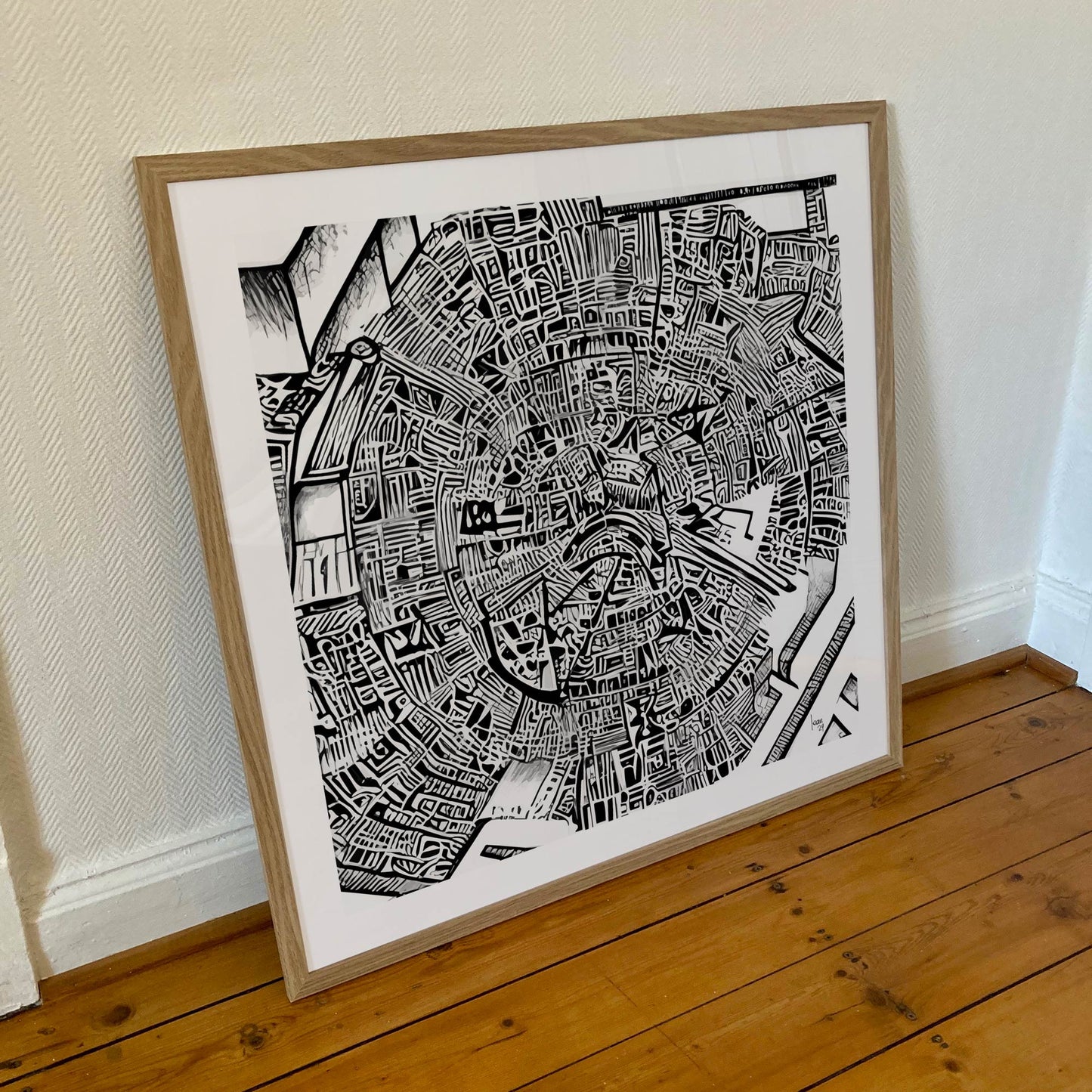 Œuvre "Matrice" encadrée au format 70×70 cm, composition graphique en noir et blanc avec un cadre en bois clair posée contre un mur blanc.