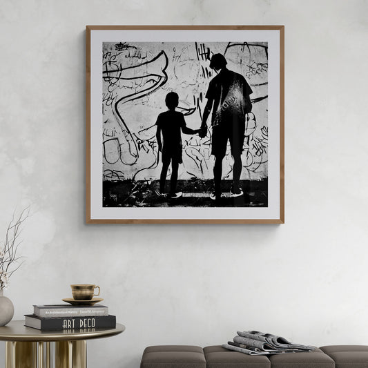 Œuvre "Paternité" sous cadre exposée dans un intérieur contemporain, tableau en noir et blanc d’un adulte et d’un enfant unis par la main, devant un mur urbain expressif.