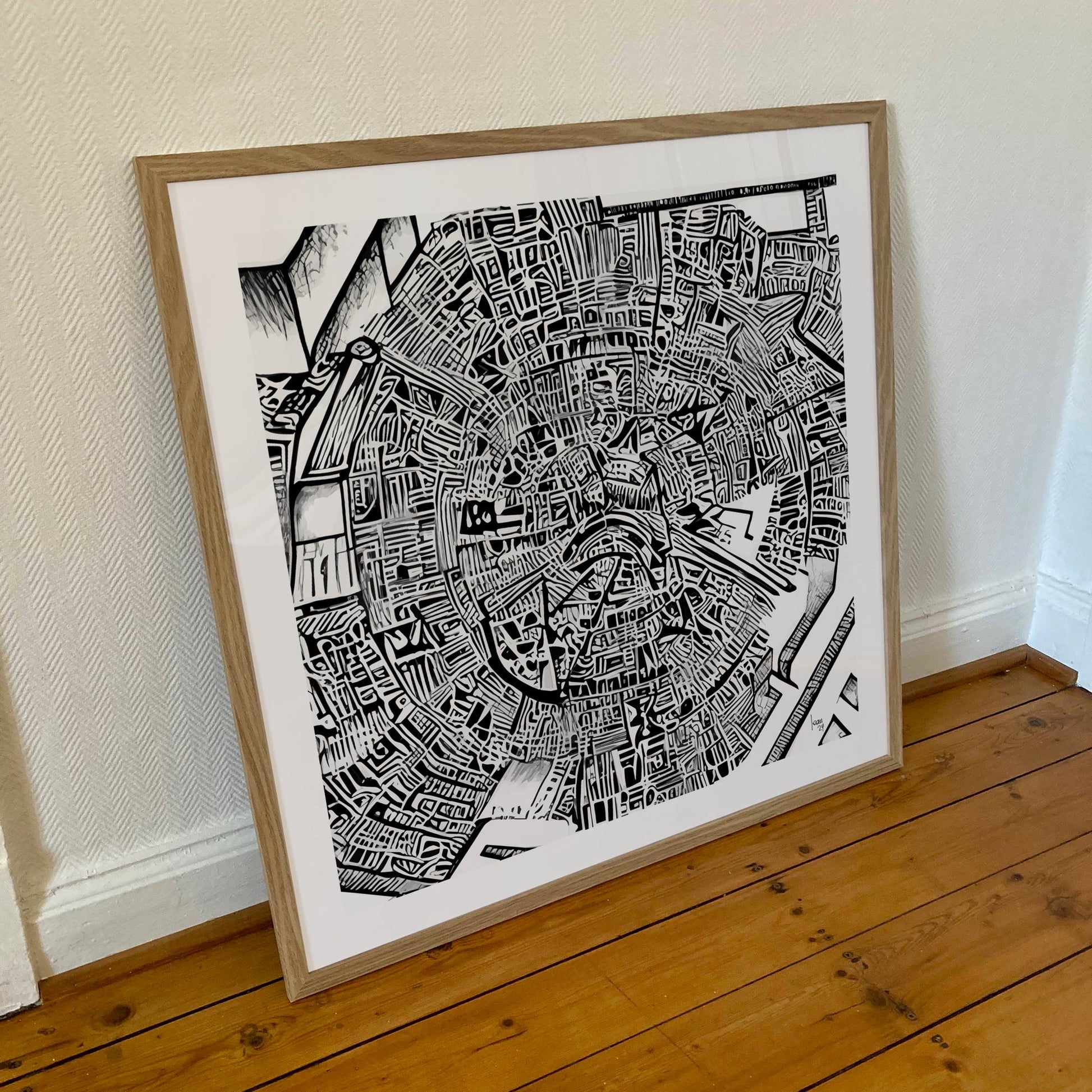 Œuvre "Matrice" encadrée au format 70×70 cm, composition graphique en noir et blanc avec un cadre en bois clair posée contre un mur blanc.